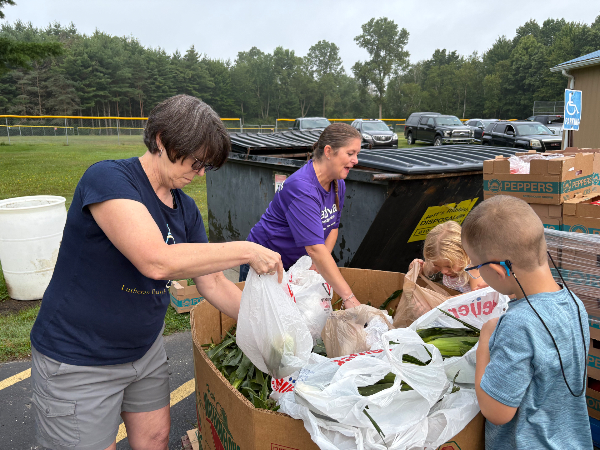 Two women and children volunteer bagging ears of corn from a bin in a parking lot.