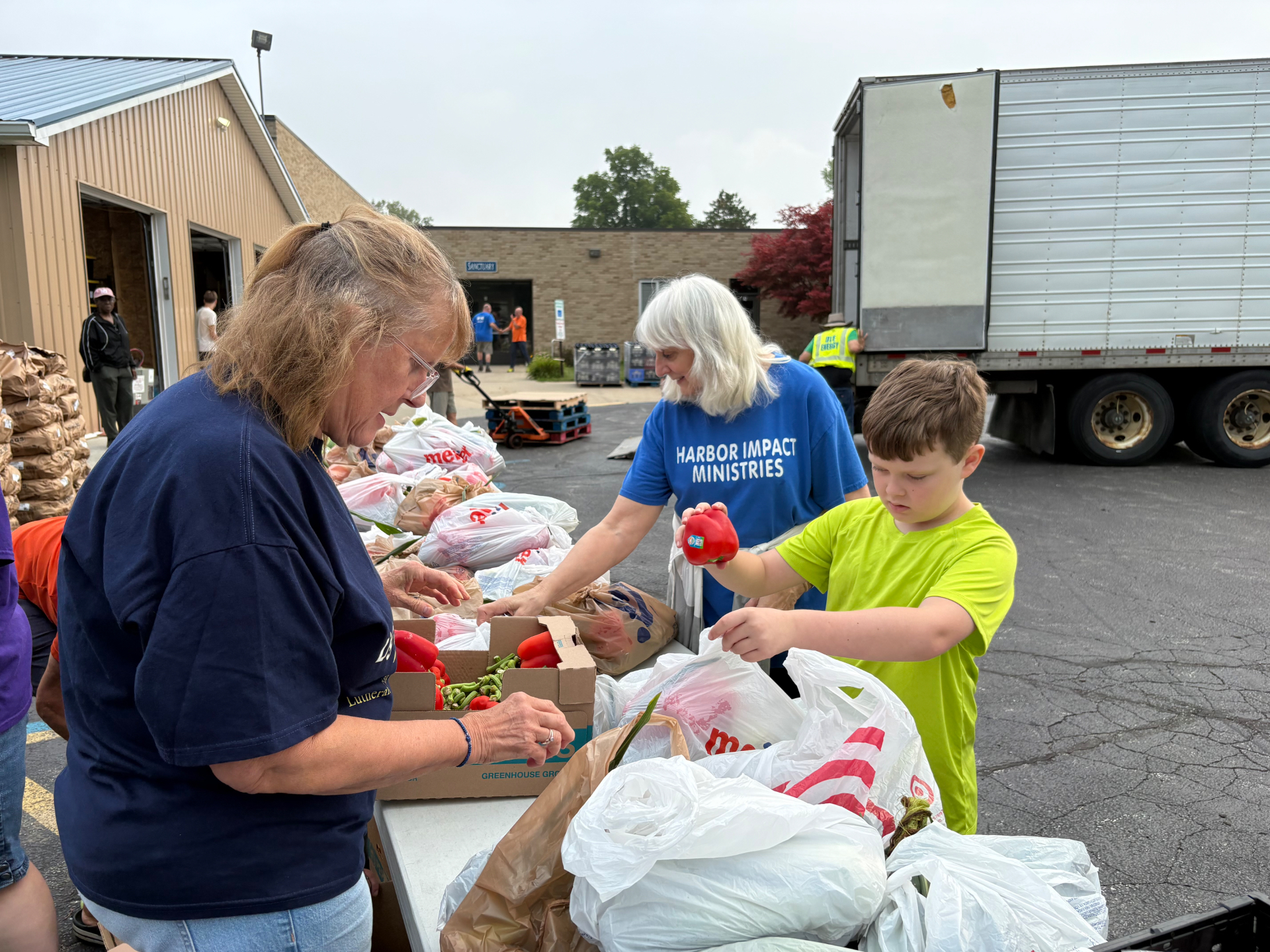 Volunteers pack fresh produce at a long outdoor table, handing peppers to a child.